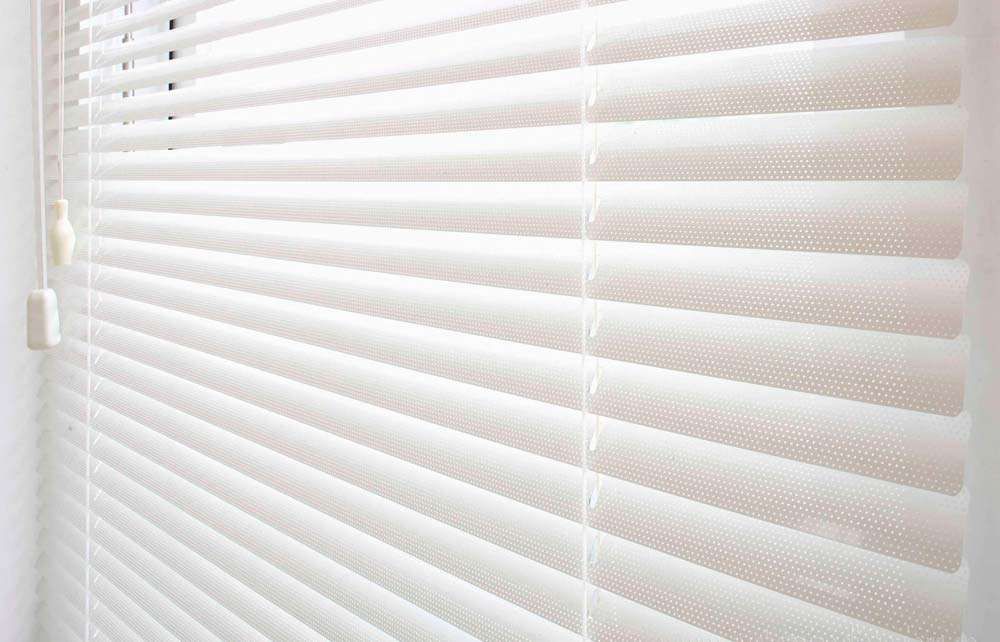 Close-up of white venetian blind slats perforated with pinholes, tilted open, with cord and wand pulls at the left