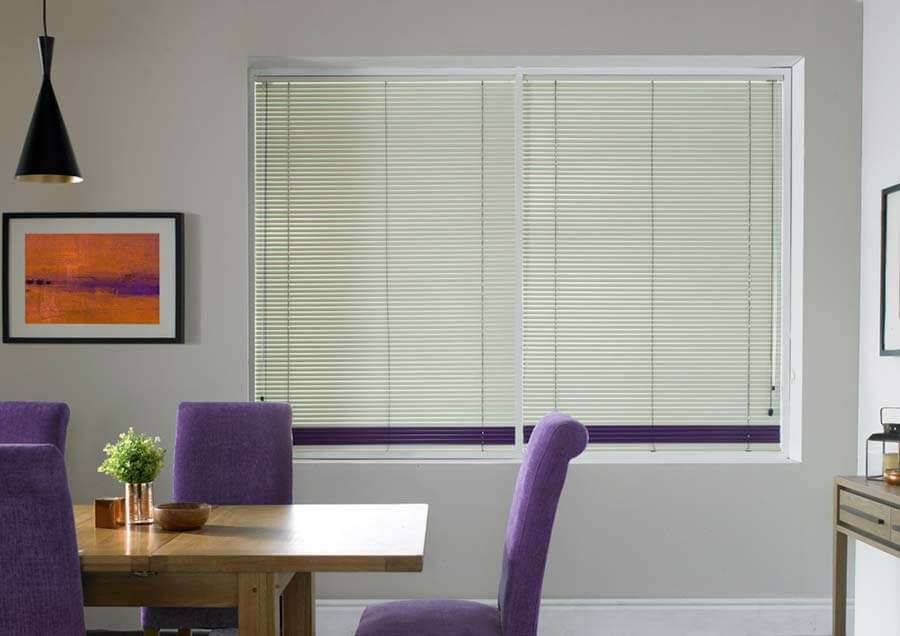 Cream aluminium venetian blinds with a purple bottom rail across a dining room window above purple upholstered chairs