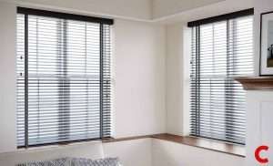 Charcoal wooden venetian blinds on two windows meeting at a corner in a white room with a grey rug