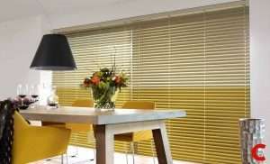 Mustard-gold venetian blinds filling a wide dining room window behind a table set with flowers and a black pendant lamp