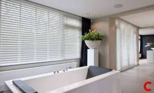White wooden venetian blinds along a bathroom window above a freestanding stone bath, dark drape at the far edge