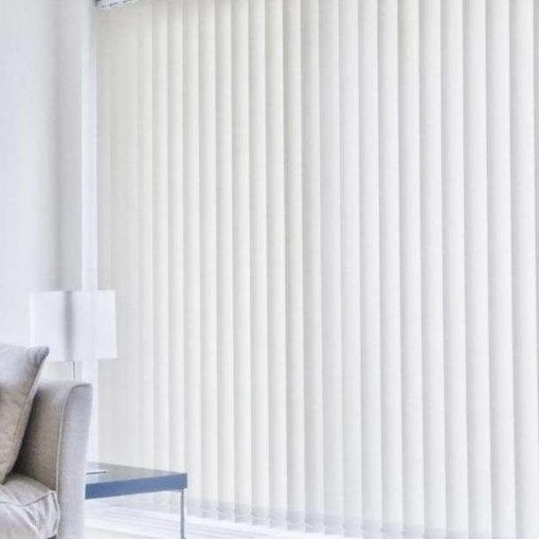 White vertical blinds closed across a full-height living room window, with a grey armchair and floor lamp alongside