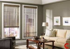 White wooden blinds on two windows in a sage-grey living room, beside a cream sofa and table lamp