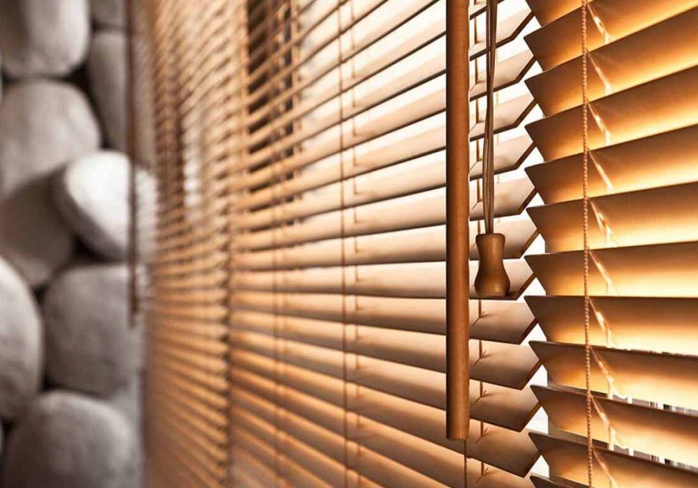 Angled close-up of warm oak wooden blind slats and their wooden cord pull, against a stacked pebble wall