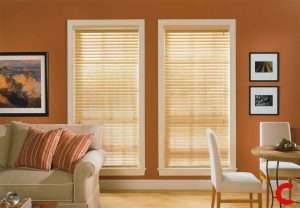 Pale golden wooden blinds on two windows in a terracotta-walled living room with a cream sofa and framed prints