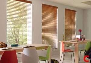 Mid-oak wooden blinds on three tall windows above a long table ringed with green and orange chairs
