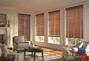 Dark walnut wooden blinds on four bay windows in a living room with a white fireplace and patterned armchairs
