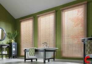 Light wooden blinds on three tall windows under a sloped ceiling in an olive-green bathroom with a freestanding bath