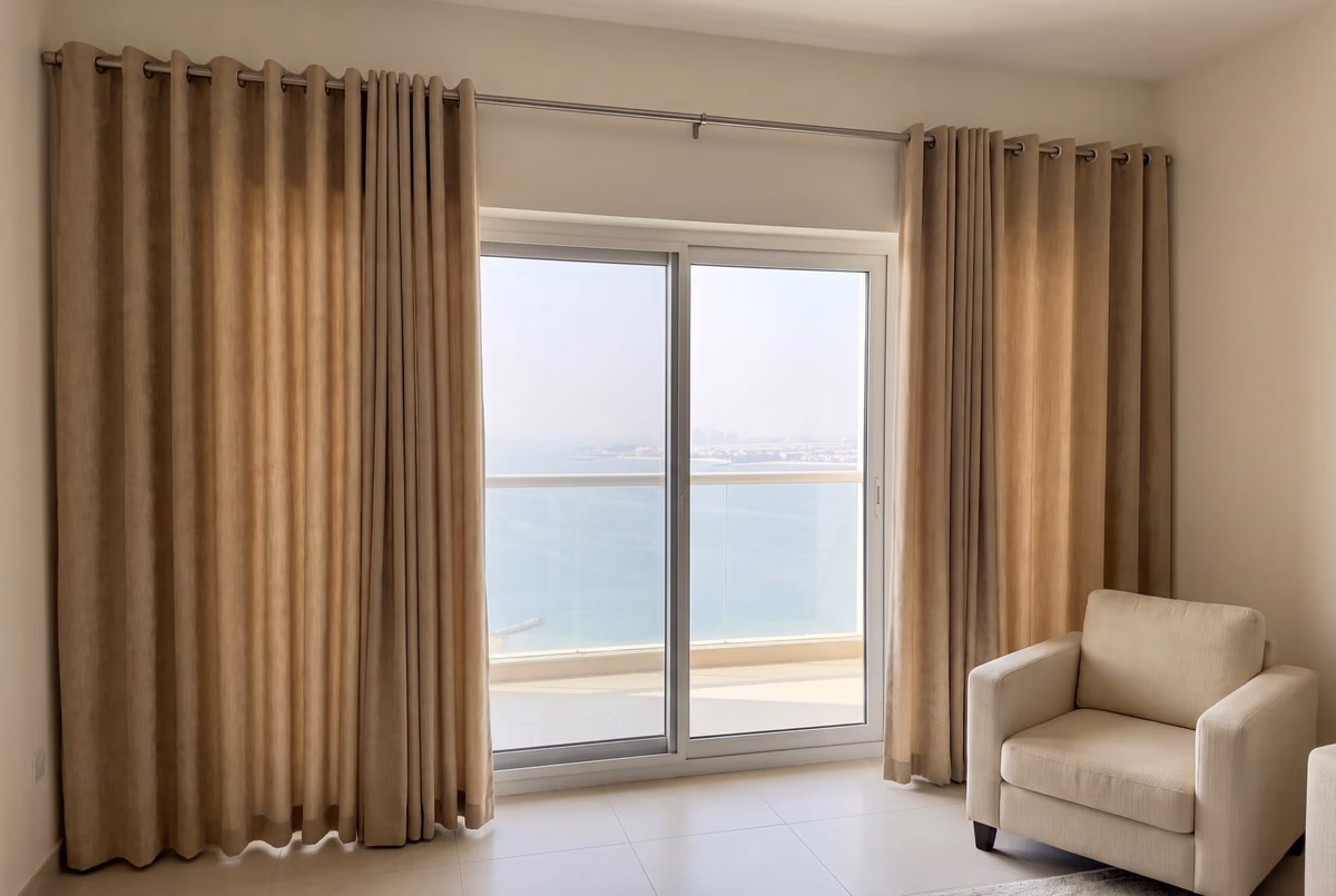 Beige eyelet curtains stacked on the wall either side of a sliding door, the pole running past the frame, the sea beyond the balcony