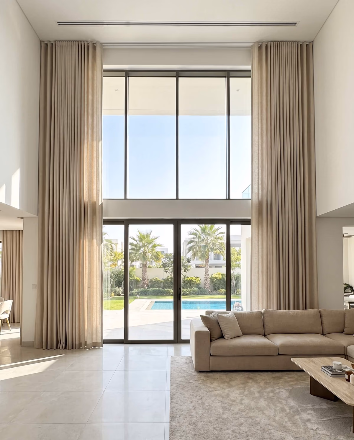 Double-height villa living room, beige wave curtains hung from the ceiling and drawn to each side of tall glass, a pool and palms outside