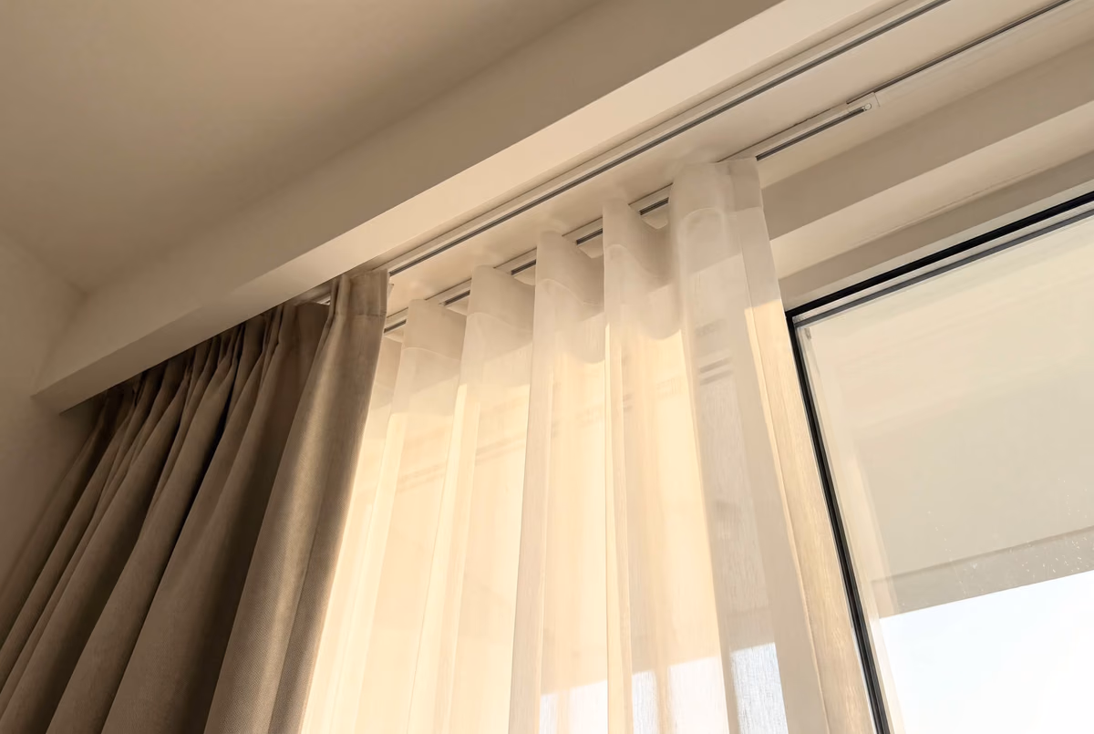 Looking up into a curtain pocket with two tracks: a beige pinch-pleat curtain on one, a white wave sheer on the other beside the glass