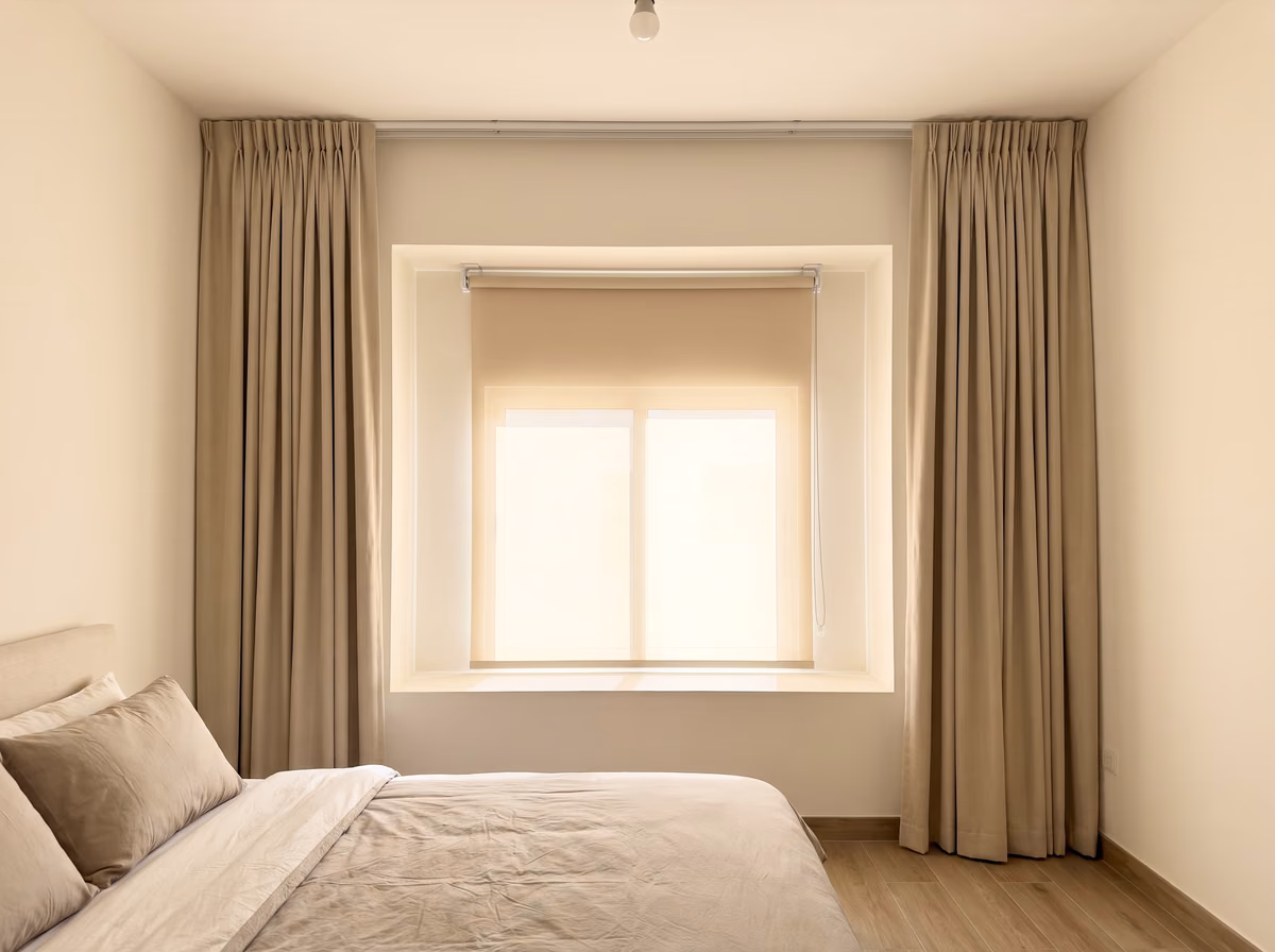 Bedroom with a beige translucent roller blind lowered inside the window recess, glowing with daylight, sand curtains drawn back either side