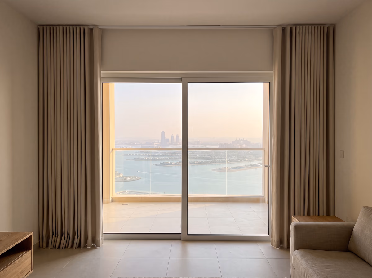 Beige ripple-fold curtains drawn back clear of a sliding balcony door in a high apartment, with the sea and islands beyond