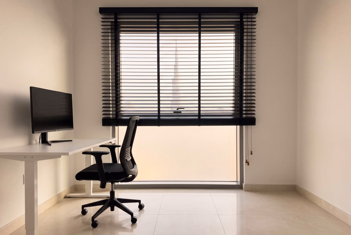 Home office with a black wooden blind raised partway over a tall window, the Burj Khalifa beyond the slats, a white desk and monitor and a black chair in front