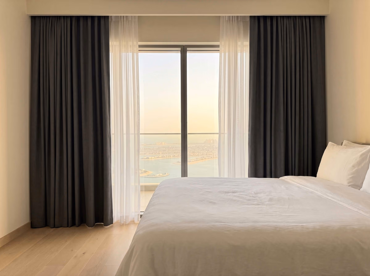 Bedroom with charcoal blackout curtains and white sheers drawn back from a balcony door, the Palm Jumeirah below at sunset, a white bed in front