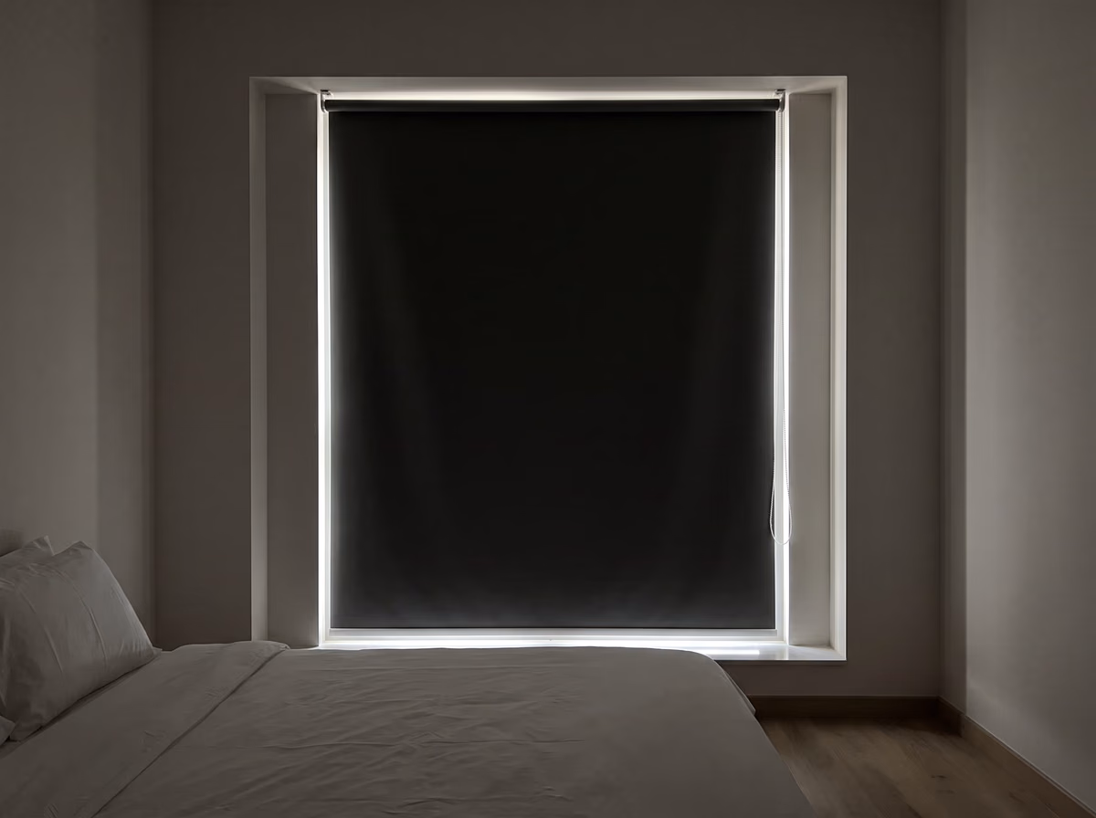 Dark bedroom in the morning, a charcoal blackout roller blind inside a deep window recess with bright lines of daylight down both sides and along the sill
