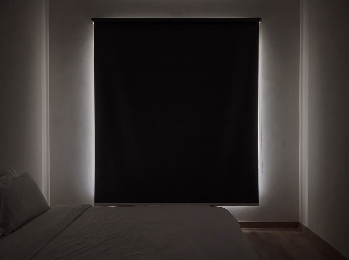 The same bedroom with a wider blackout roller blind fixed on the wall over the window, only a soft glow showing round its edges