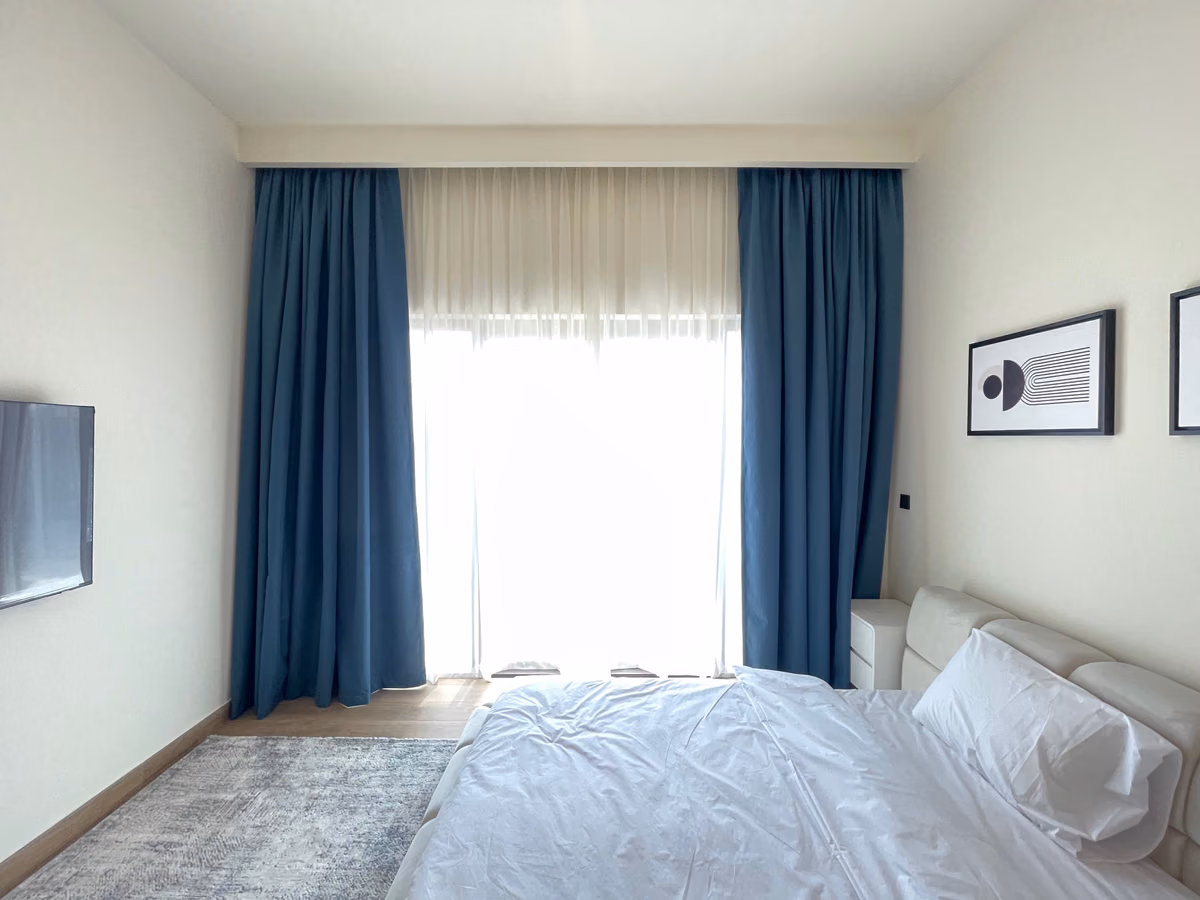Blue blackout curtains either side of a sunlit bedroom window with a white sheer drawn, a bed with a padded headboard to the right