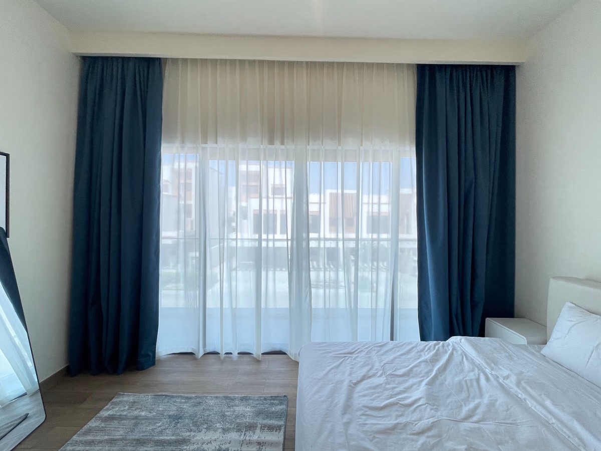 Blue blackout curtains stacked either side of a bedroom's balcony doors, a white sheer drawn across and houses beyond