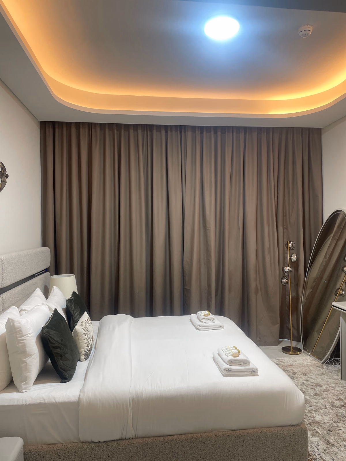 Brown blackout curtains drawn across a bedroom window from the ceiling pocket to the floor, behind a made bed with folded towels