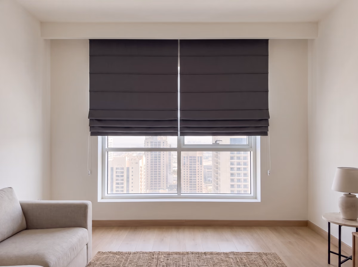 Two charcoal blackout roman blinds hung from the ceiling over a tower living room window, folds gathered at their hems above clear glass