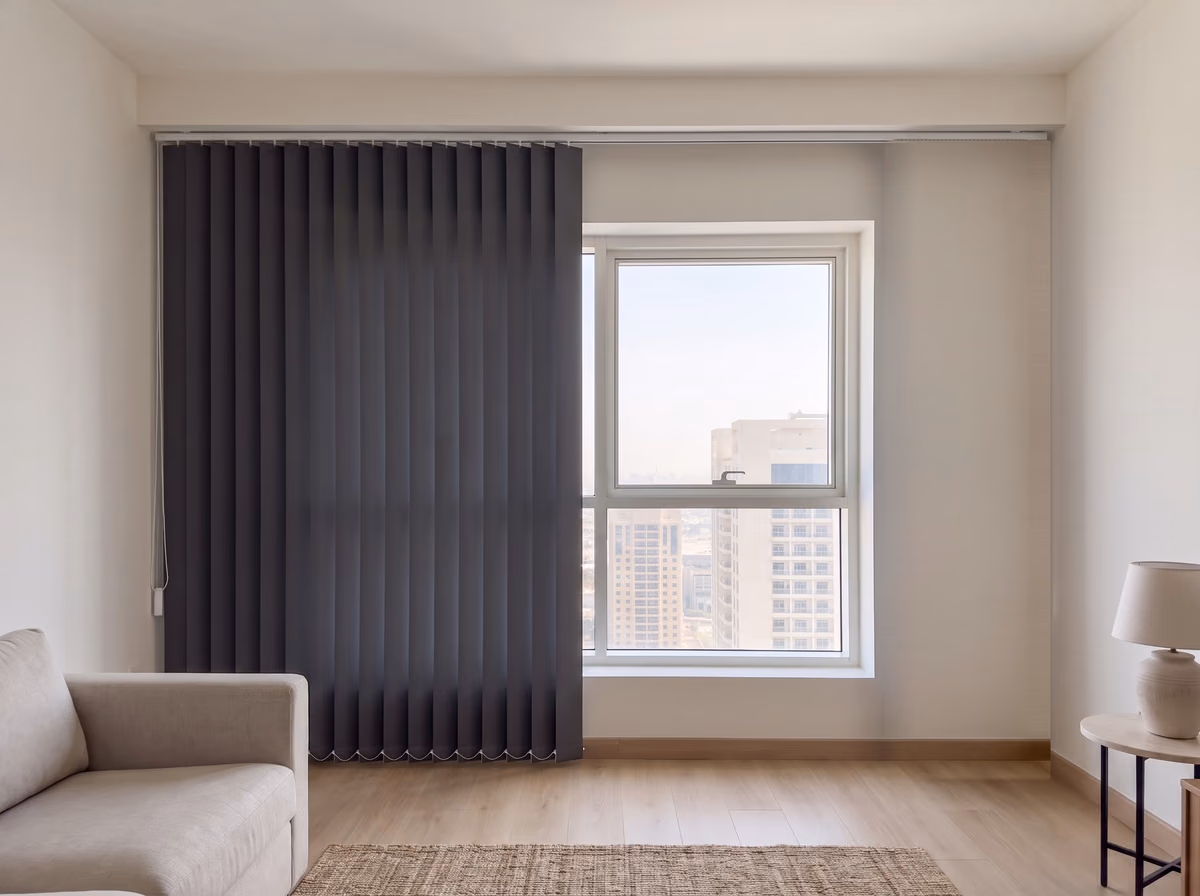 Charcoal blackout vertical blind drawn two-thirds across a tower living room window from the left, the right of the glass clear