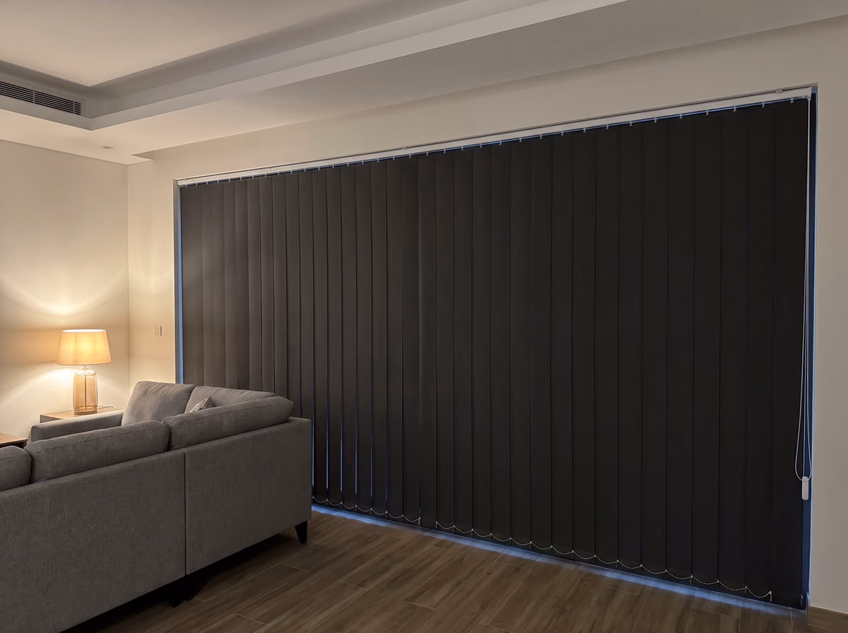 Charcoal blackout vertical blind closed across a wide villa window in the evening, a lamp lit beside a grey corner sofa