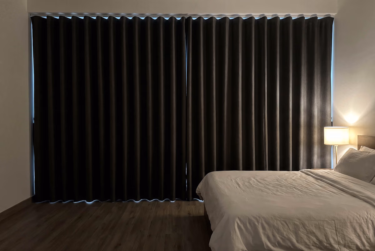 Bedroom at dusk with charcoal blackout wave curtains closed across a wide window, a thin line of light along the top and sides, a bedside lamp lit