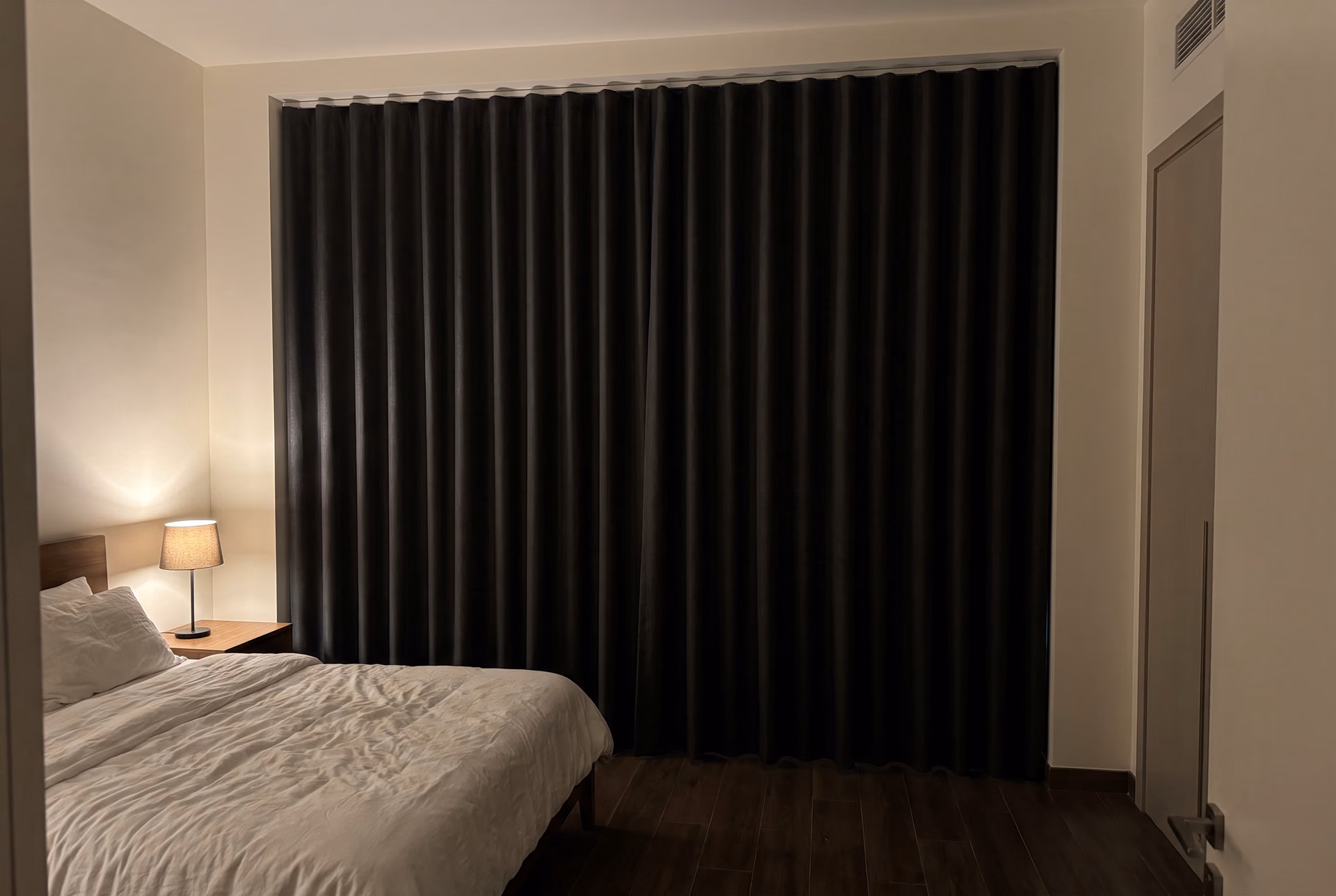 Charcoal blackout curtains in a wave heading closed across a bedroom window from a ceiling track, the room dark but for one bedside lamp