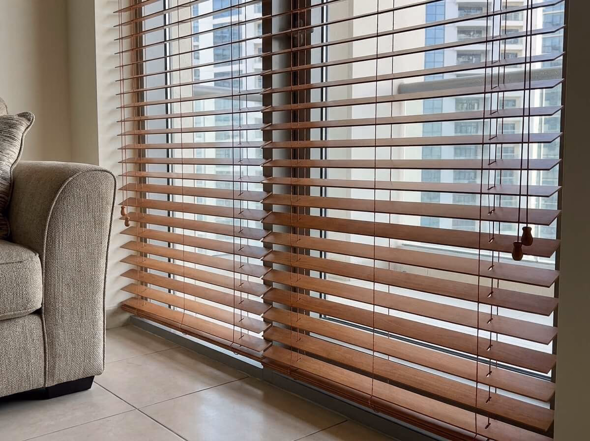 Close-up of cherry-brown wooden blinds tilted open at an apartment window, the ladder cords and wooden cord pulls showing and residential towers beyond the glass