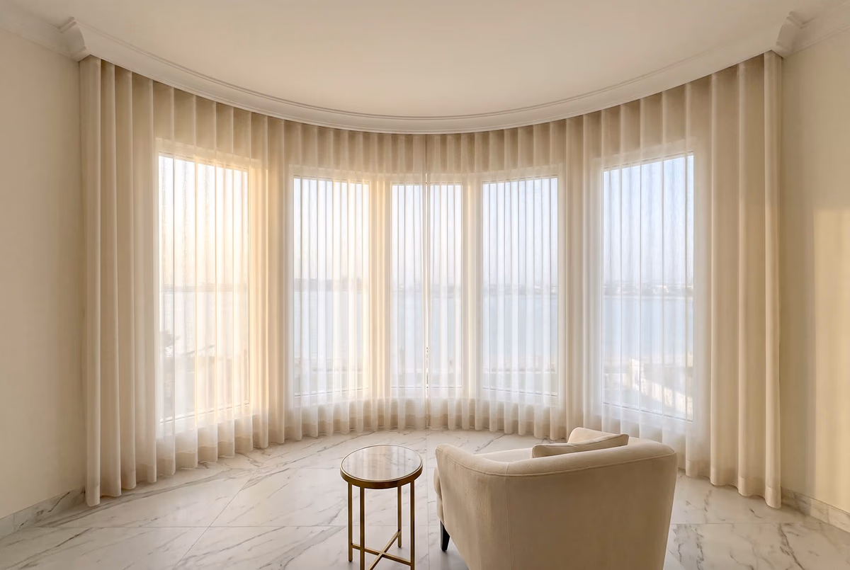 Curved bay of five windows with cream sheer wave curtains following a curved ceiling track, the sea pale beyond, an armchair in front
