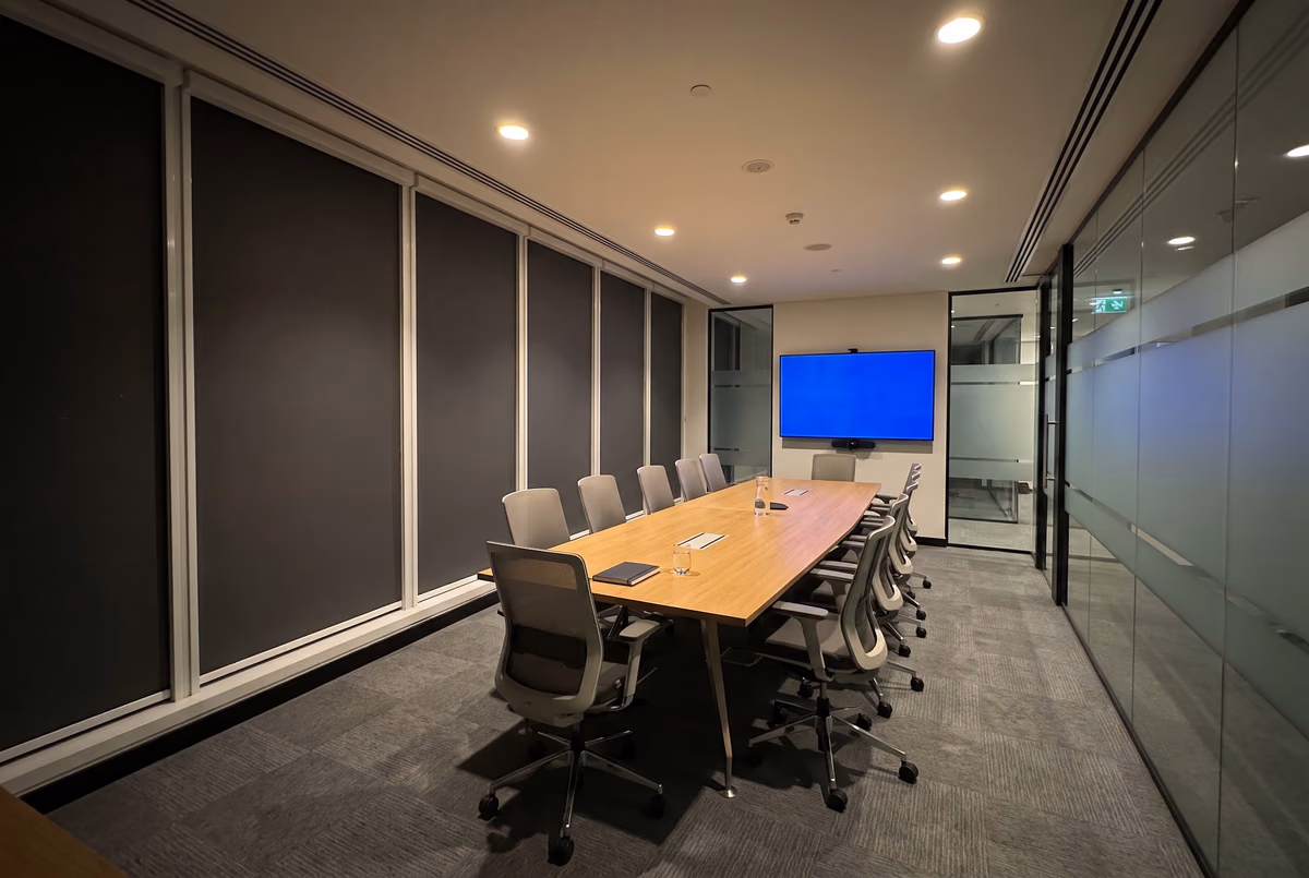Meeting room with dark grey blackout roller blinds down along the window wall, a long table and a wall screen