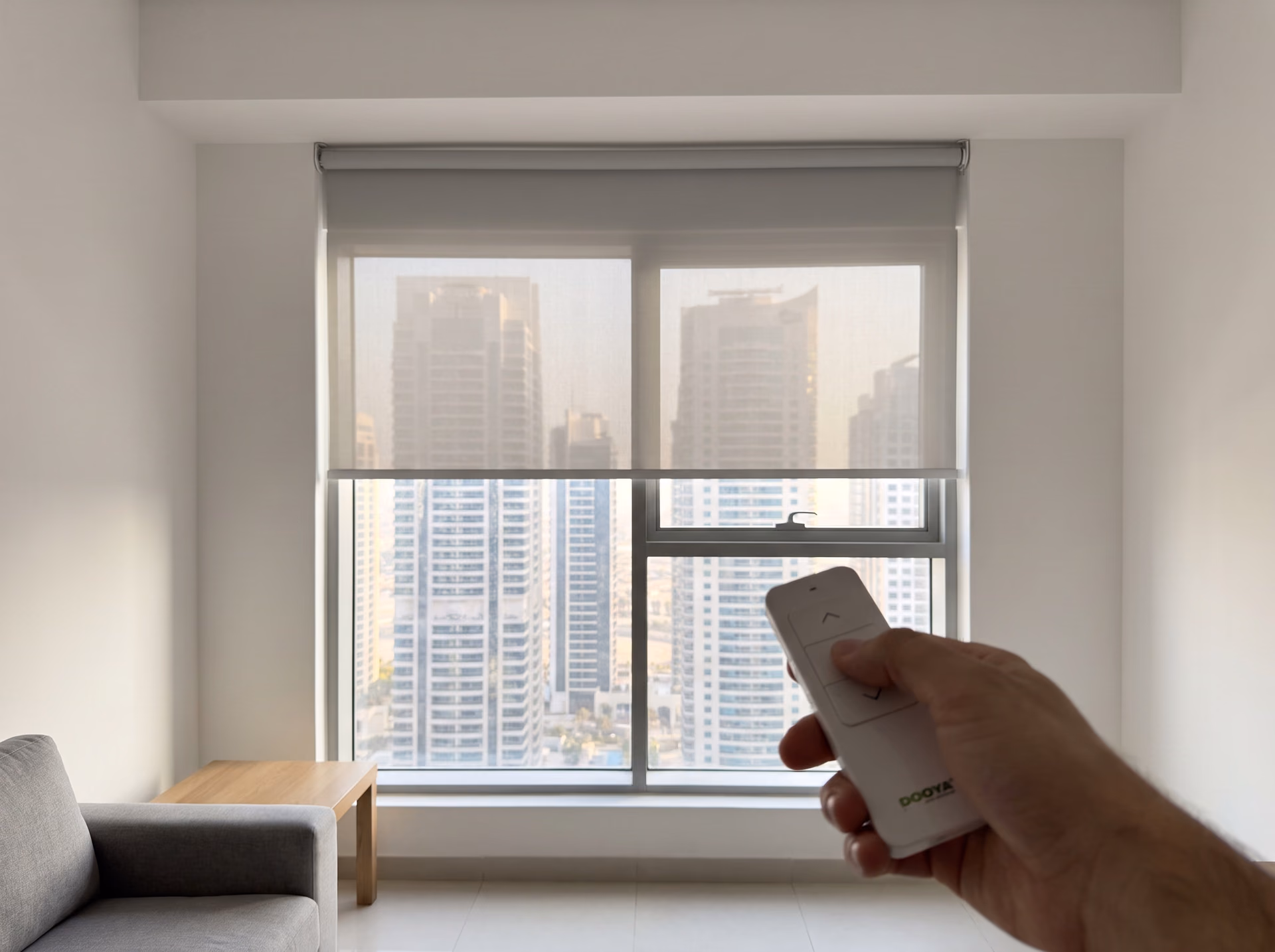 A hand pointing a white Dooya remote at a light grey sunscreen roller blind half lowered over tower windows, high-rise towers beyond