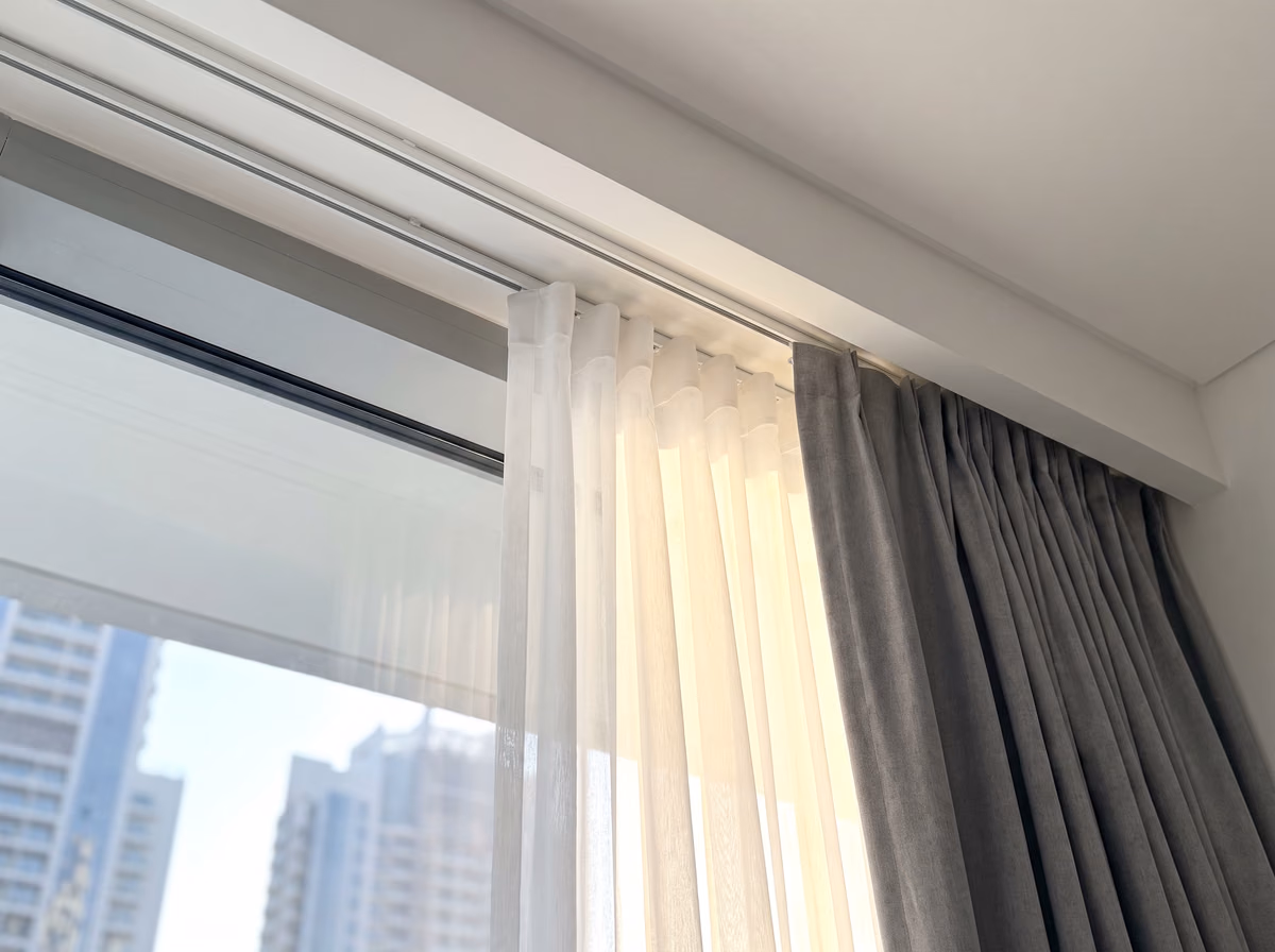 A white sheer and a grey curtain hanging from two white tracks in a ceiling recess above floor-to-ceiling glass, towers beyond