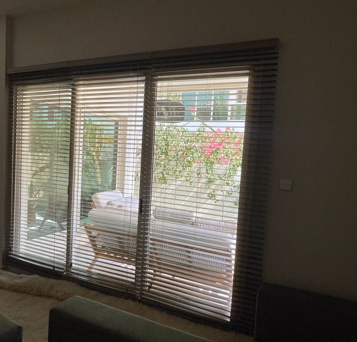 Three faux wood blinds hung above the frame of a villa's triple glass door, seen from the side of the living room, the slats tilted open onto a garden terrace