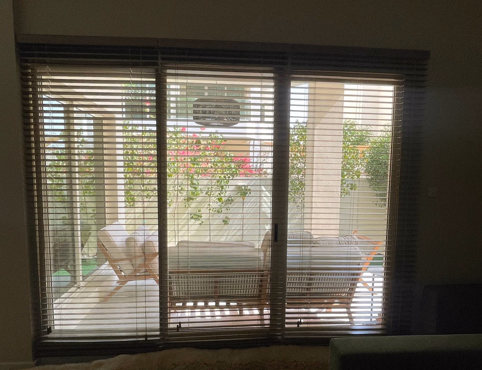 Three dark faux wood blinds with their slats tilted open across a villa's triple glass door, garden chairs and bougainvillea on the terrace beyond