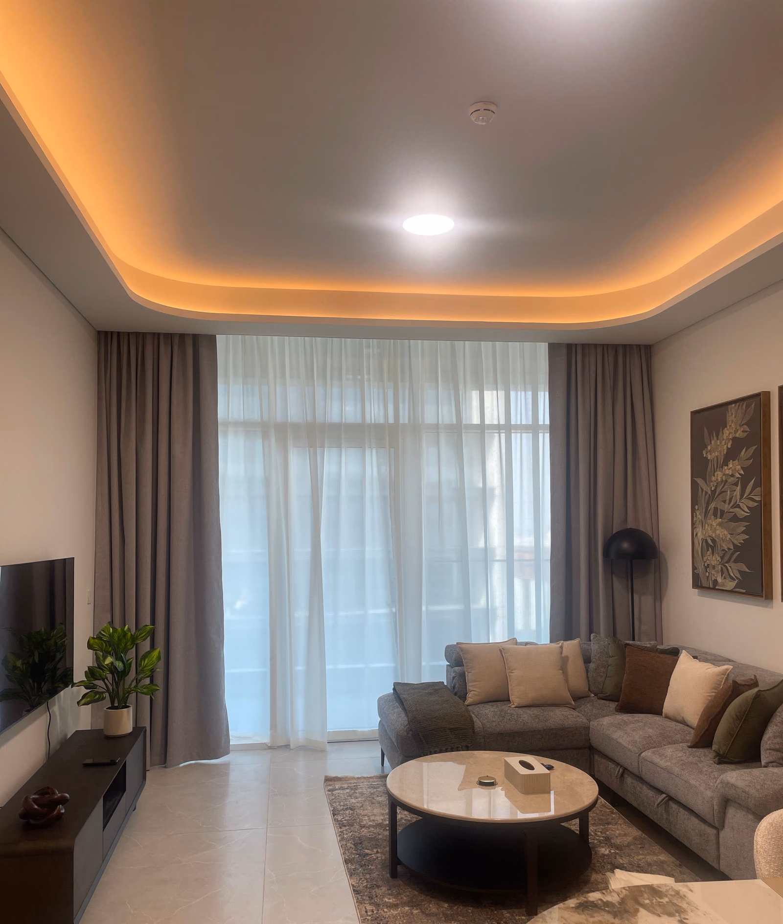 Grey blackout curtains stacked either side of a floor-to-ceiling window in an apartment living room, a white sheer drawn across the glass and a grey corner sofa in front