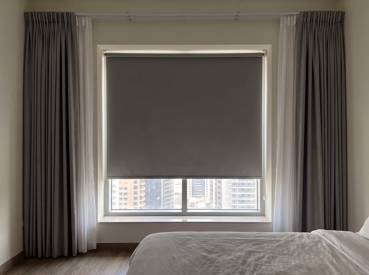 Bedroom window with a grey blackout roller blind lowered most of the way inside the recess, white sheers and grey curtains drawn back