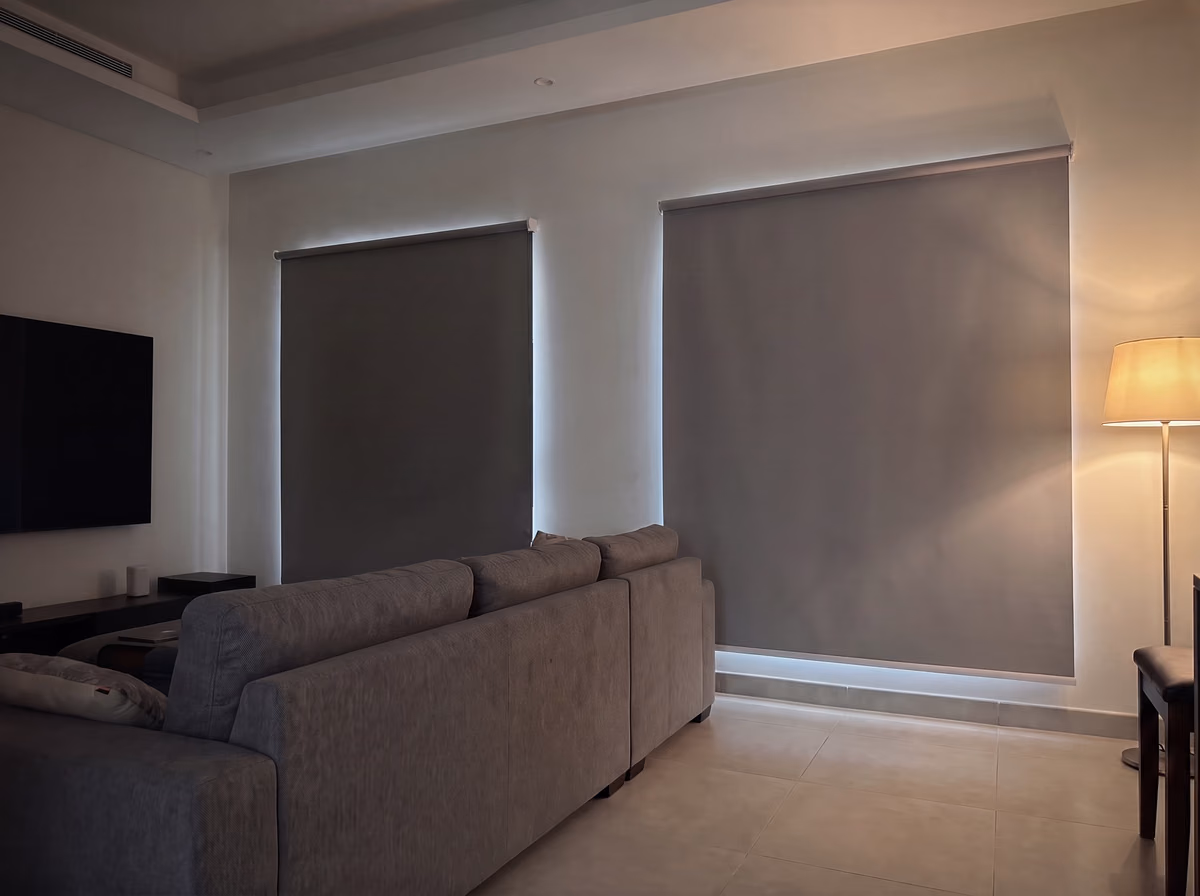 Media room with two grey blackout roller blinds lowered over the windows behind a grey sofa, no chains, a soft glow at their edges