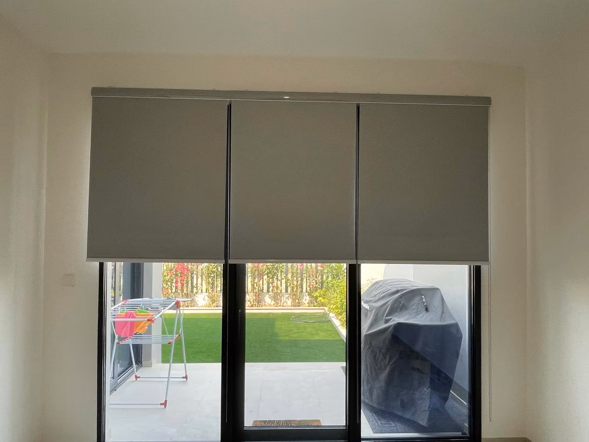 Three grey blackout roller blinds half lowered over triple glass doors in an empty room, a lawn and covered terrace visible below them