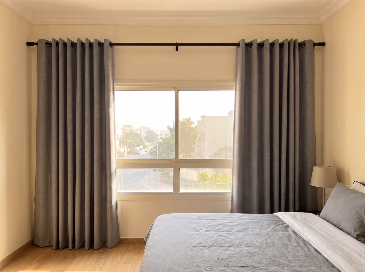 Grey eyelet curtains on a matte black metal pole, drawn back either side of a bedroom window over a villa street, a bed beside it