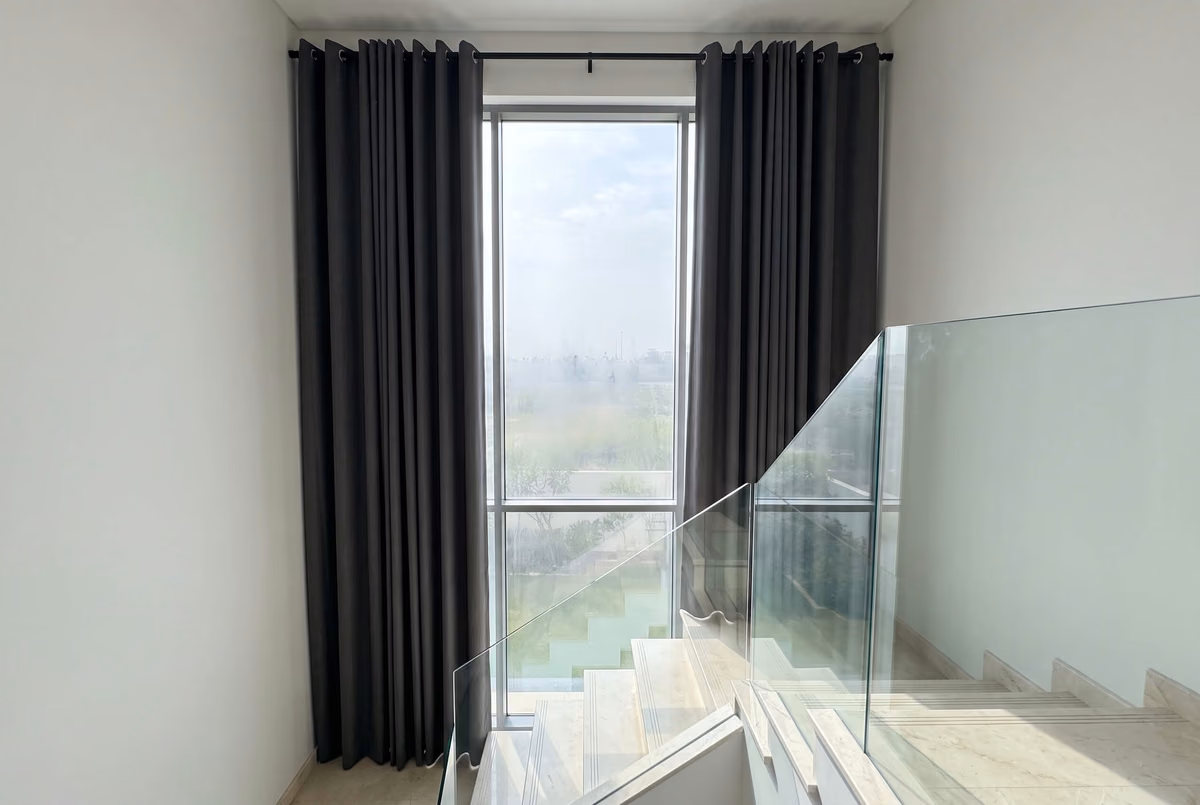 Charcoal eyelet curtains on a black pole framing a tall villa stairwell window, a glass balustrade and marble stairs below