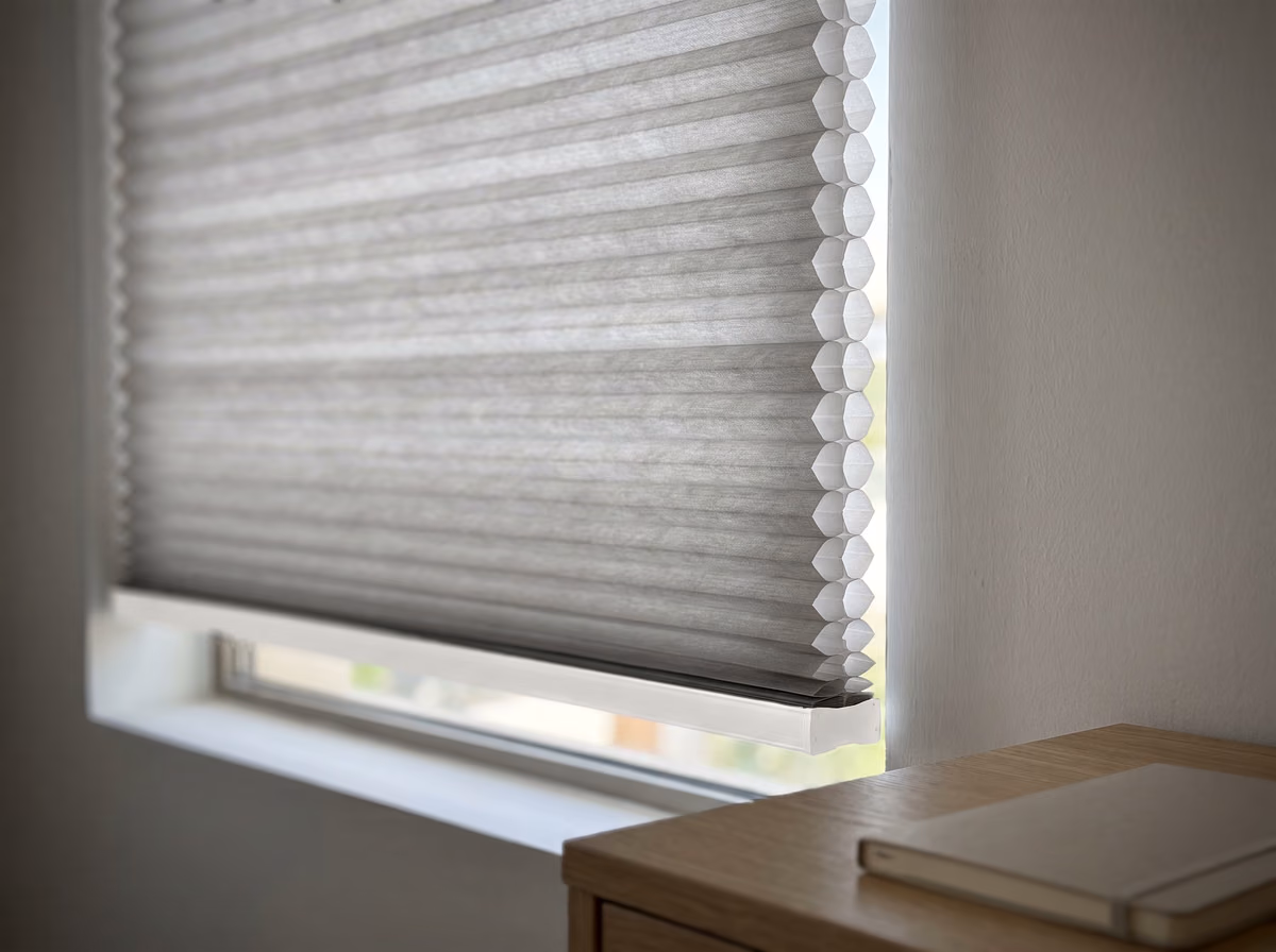 Close view of a light grey honeycomb blind inside a window recess, its cut edge showing a column of six-sided cells lit by daylight