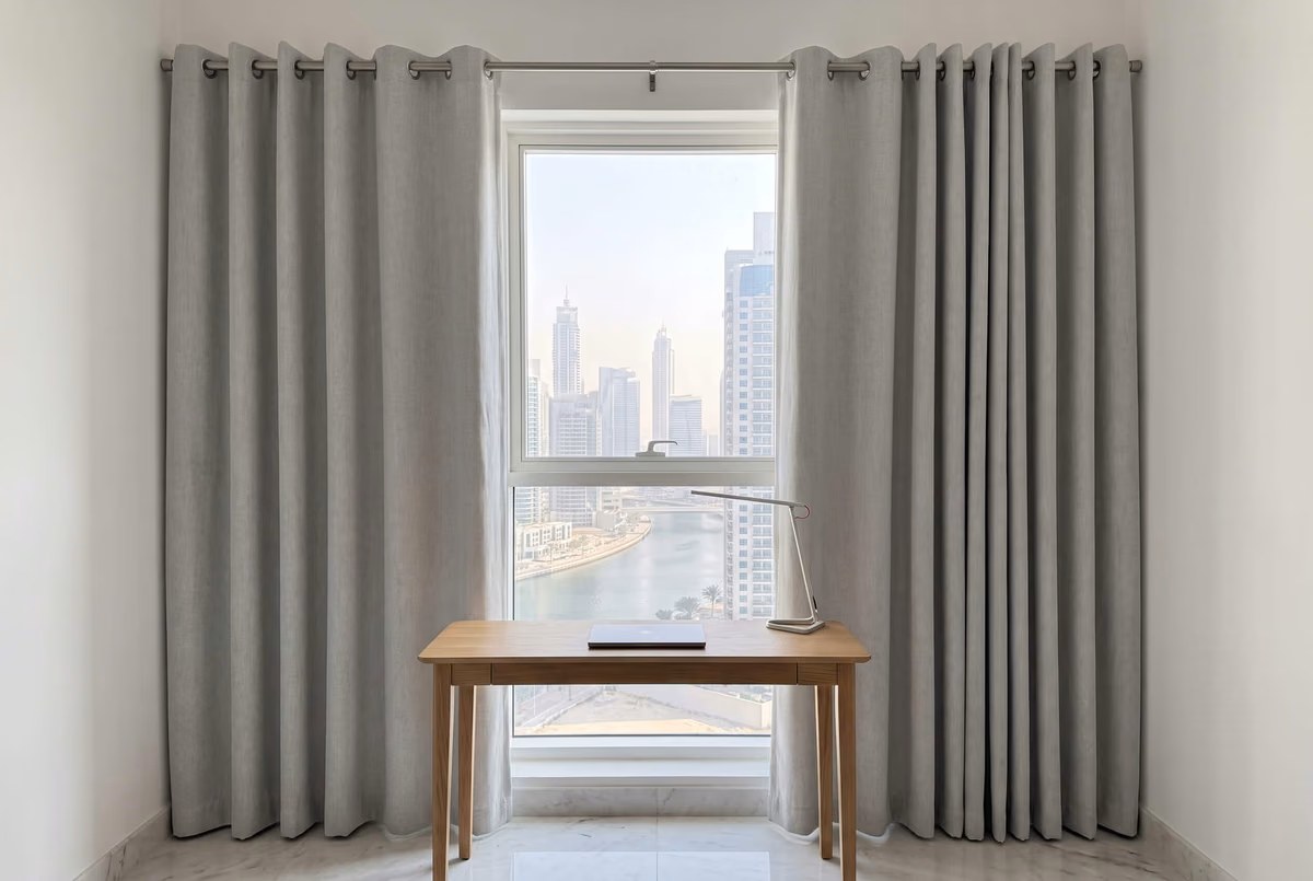 Pale grey eyelet curtains on a brushed-steel pole either side of a study window, a desk in front and Dubai Marina beyond