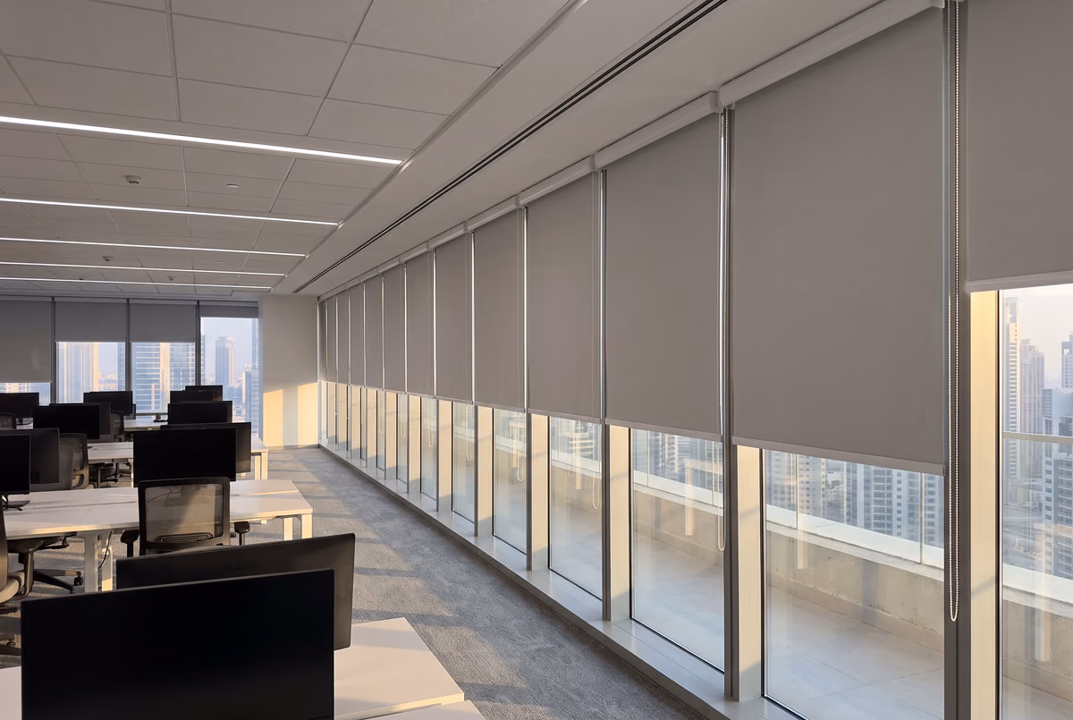 Grey roller blinds lowered part way along a long run of tower windows above an open-plan floor of desks