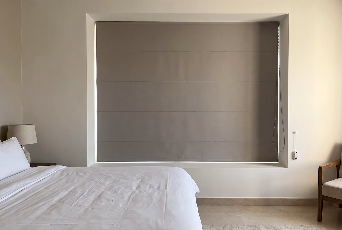 Grey roman blind lowered inside a deep window recess above a bed, its chain hanging at the right