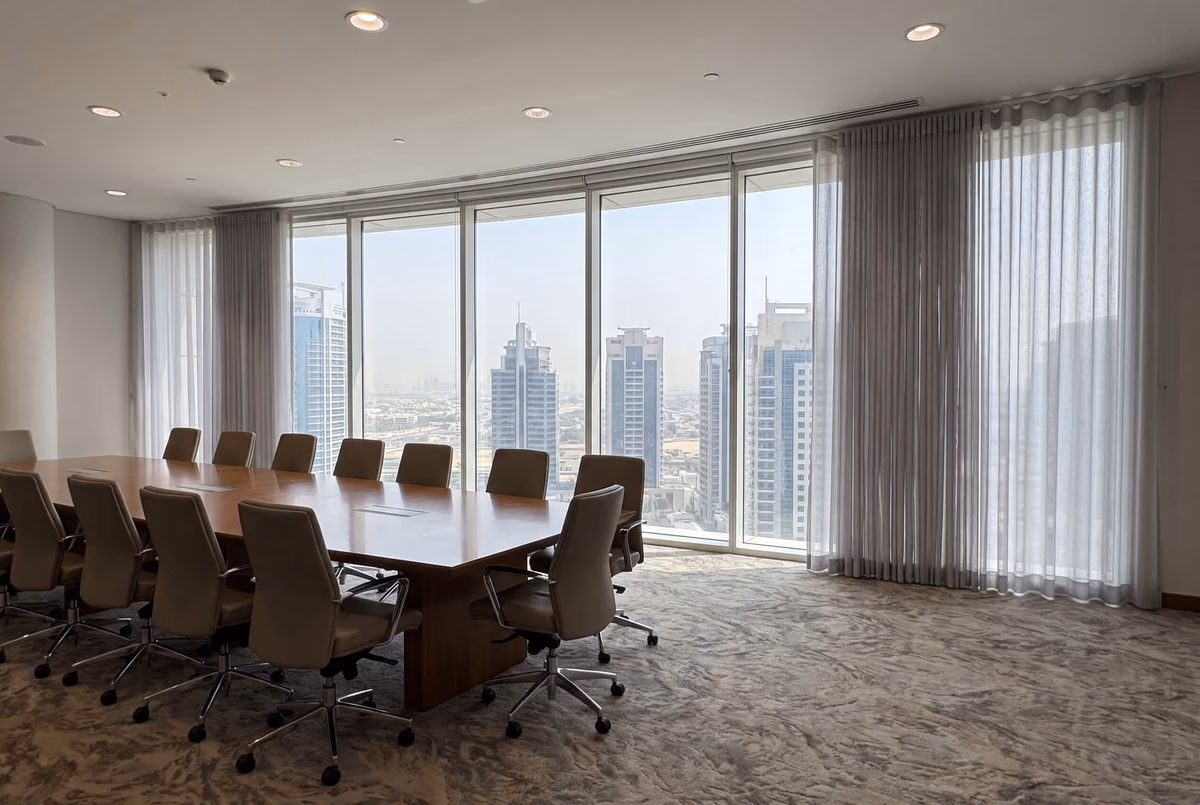 Boardroom with light grey sheer wave curtains drawn to each side of floor-to-ceiling glass, towers outside and a long table with leather chairs