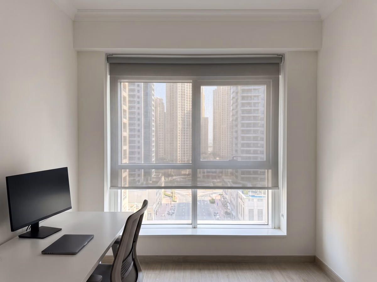 Home office with a grey sunscreen roller blind lowered over a tower window behind a desk and a monitor, the street below in clear glass