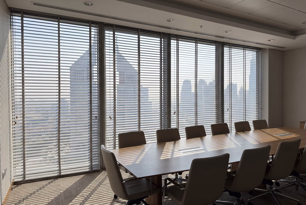 Boardroom with grey wooden blinds along floor-to-ceiling glass, towers beyond the tilted slats and stripes of sunlight across a long table