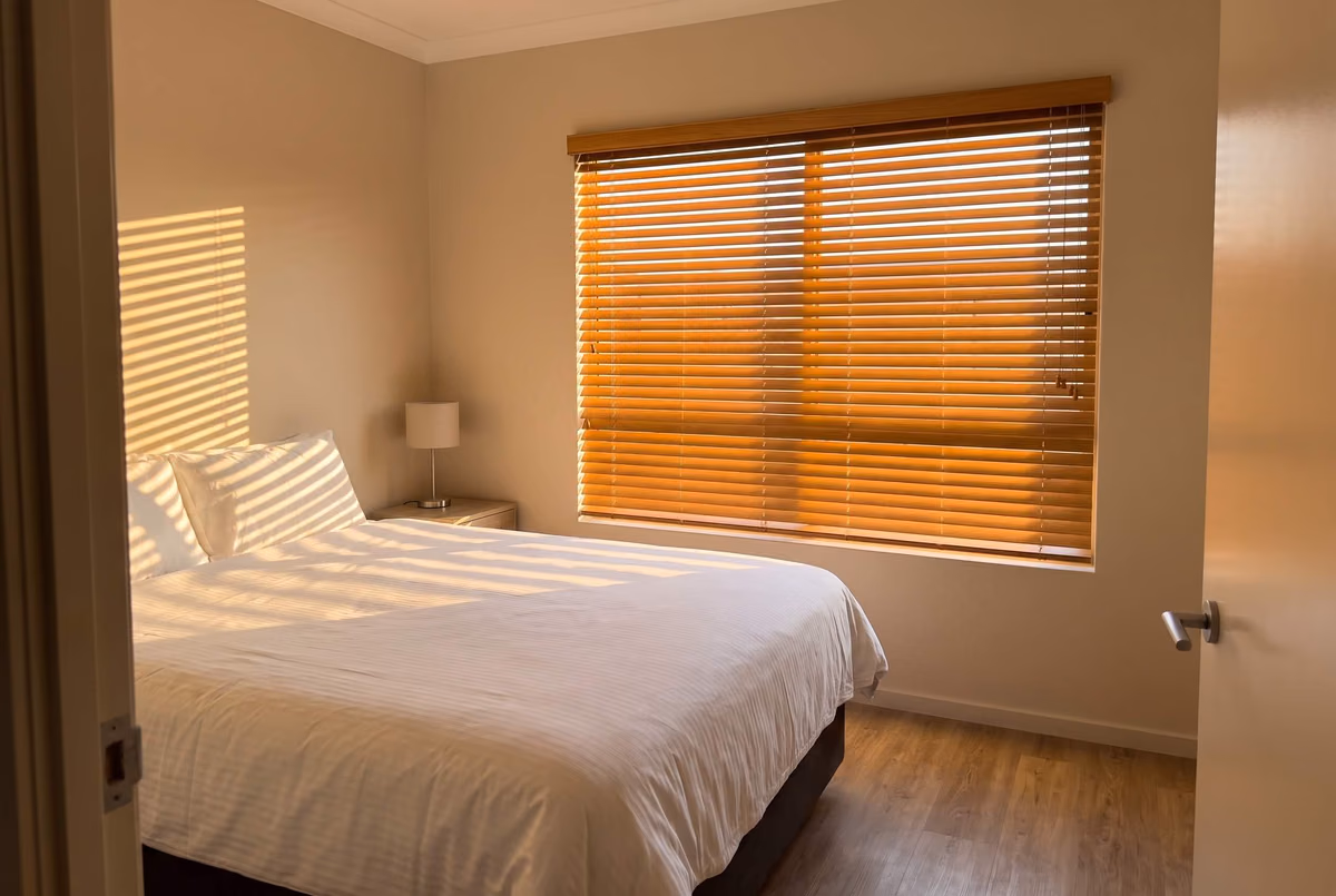 Bedroom with a honey wooden blind lowered over a wide window in low evening sun, light through the slats striping the wall and the white bedding
